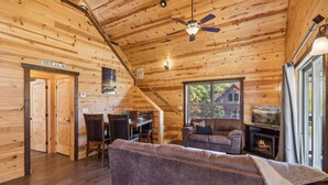 A wooden cabin interior with a sofa, fireplace, dining area, and a staircase leading to a loft.