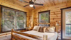 A wooden cabin bedroom with a large bed, a ceiling fan, and a window with blinds.