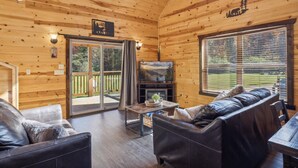 A cozy cabin interior with wooden walls, a fireplace, leather sofas, and a large window offering a view of the outdoors.