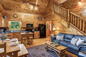 Cozy cabin interior with a wooden staircase, dining area, and leather sofa, set amidst a natural view.