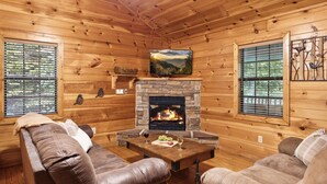 Un interior de cabaña acogedor, con chimenea de piedra, sofás de cuero y un televisor montado encima.