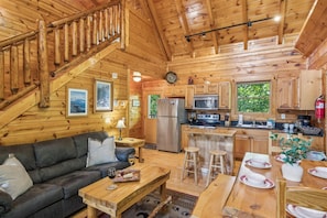 Ein gemütliches Blockhaus-Interieur mit einer Holztreppe, einem Wohnbereich mit Sofa und Couchtisch, einer Küche mit modernen Geräten und einem Essbereich mit Tisch für vier Personen.
