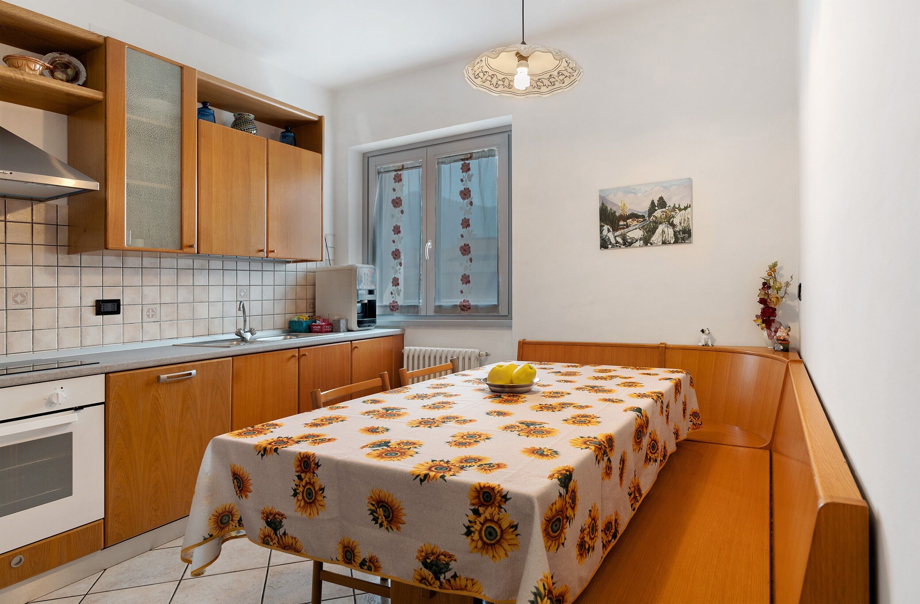 Een keuken met houten kastjes, een tafel met een tafelkleed met zonnebloemen, een fruitschaal en een raam met bloemen gordijnen.