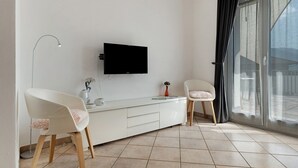 A modern room with a flat-screen TV, a white cabinet, two chairs, a floor lamp, and a large sliding glass door.