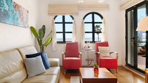 A living room with a beige sofa, two red armchairs, a wooden coffee table, and a large window with a view. - Luzias Apartment with Sea View, Wi-Fi & Balcony (Funchal)