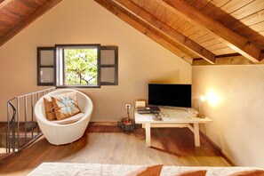 A room with a sloped ceiling, a wicker chair, a table with a flat-screen TV, and a window with shutters.
