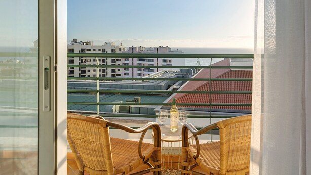 A balcony with wicker chairs and a table, overlooking a coastal cityscape with buildings and a clear sky. - Holiday Apartment Oliveira Atlantic View with Sea View, Wi-Fi & Balcony (Funchal)