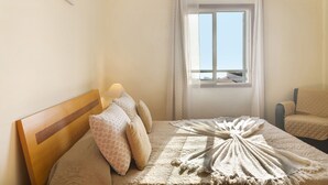 A bedroom with a wooden headboard, a bed with patterned pillows, a quilted blanket, a chair, and a window with curtains. - Holiday Apartment Oliveira Atlantic View with Sea View, Wi-Fi & Balcony (Funchal)