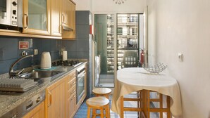 A compact kitchen with a sink, stove, and wooden cabinets. There is a dining table with a white tablecloth and two stools. - Holiday Apartment Oliveira Atlantic View with Sea View, Wi-Fi & Balcony (Funchal)