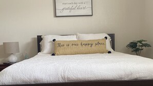 A neatly made bed with a white comforter and a decorative pillow stating "This is our happy place." A framed inspirational quote hangs above the bed.