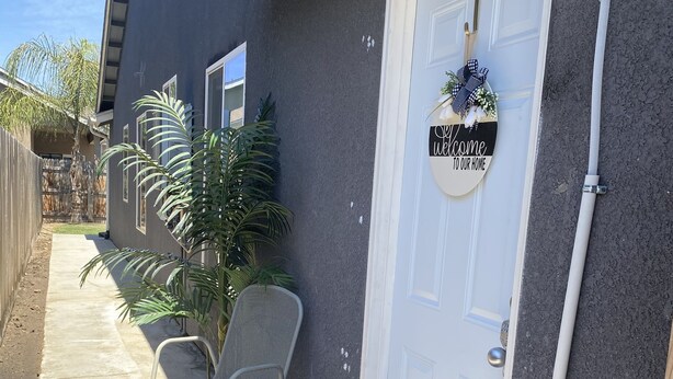 A house exterior with a dark gray exterior, a white door with a welcome sign, a potted plant, and a chair on a concrete porch.