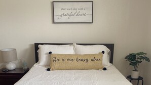 A neatly made bed with a white textured bedspread, two pillows, and a decorative pillow with a message. There is a framed inspirational quote on the wall above the bed.