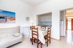A dining area with a table set for four, a sofa, a kitchen in the background, and a beach scene on the wall.