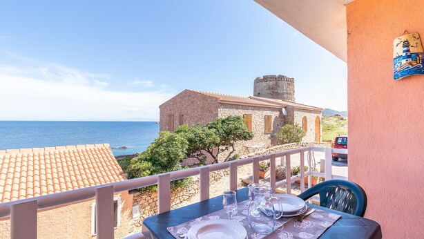 A balcony with a table set for dining, overlooking a coastal view with a stone building and a tower. - Apartment "Vista Corsica" on Isola Rossa with Sea View & Terrace (Isola Rossa)
