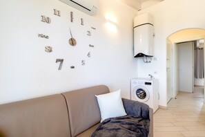 Quarto com sofá bege, relógio na parede, máquina de lavar e corredor que dá para outro cômodo.