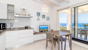 A modern kitchen with white cabinets, a dining area with wicker chairs, a TV, and a view of the sea through a sliding glass door. - Apartment "Vista Bocche di Bonifacio" with Outdoor Pool (Isola Rossa)