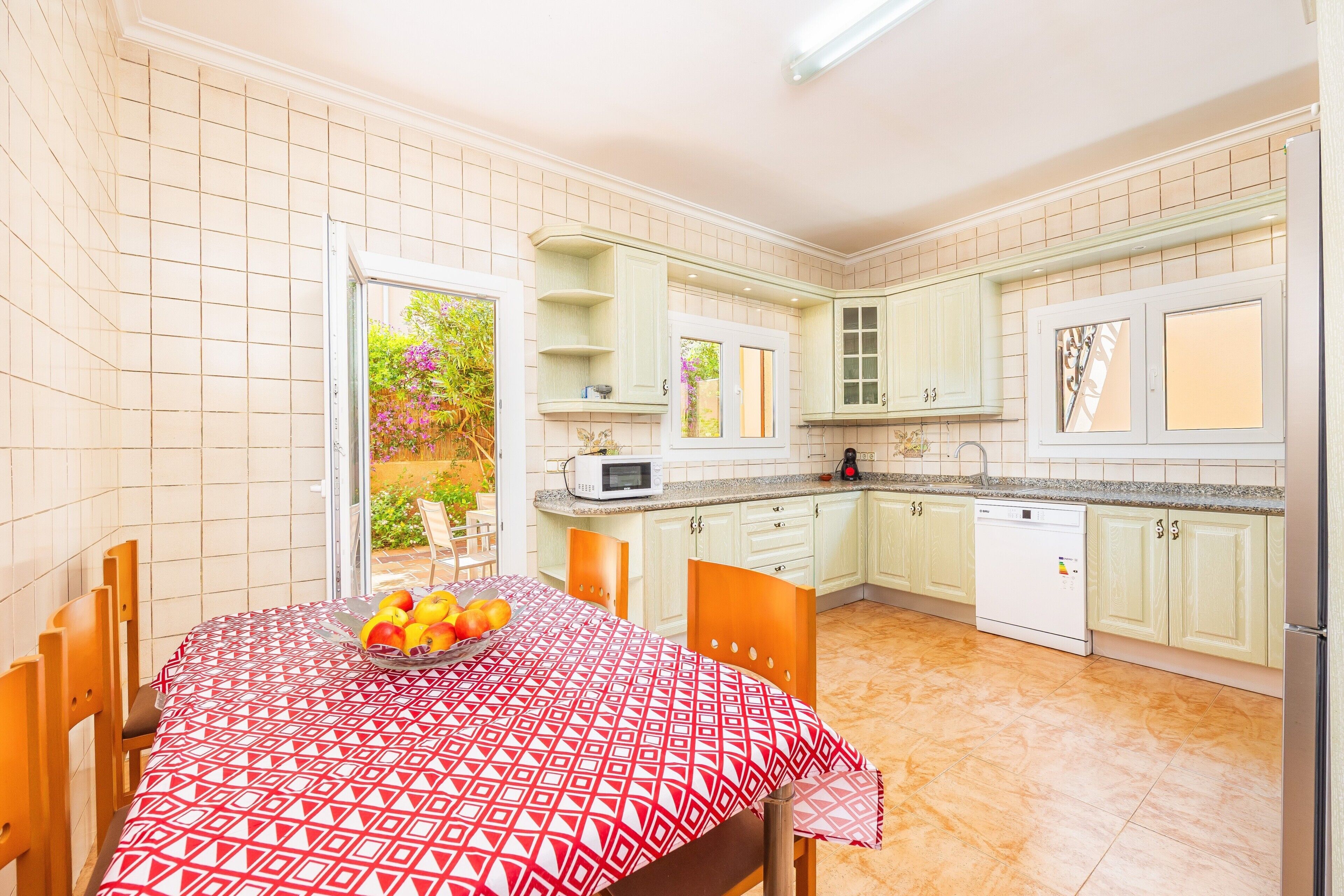 Een keuken met witte kastjes, een eettafel met een tafelkleed met een patroon, en een fruitschaal.