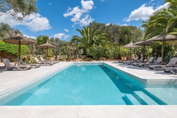 Una piscina con sillas de descanso y sombrillas de paja, rodeada de plantas y árboles tropicales.