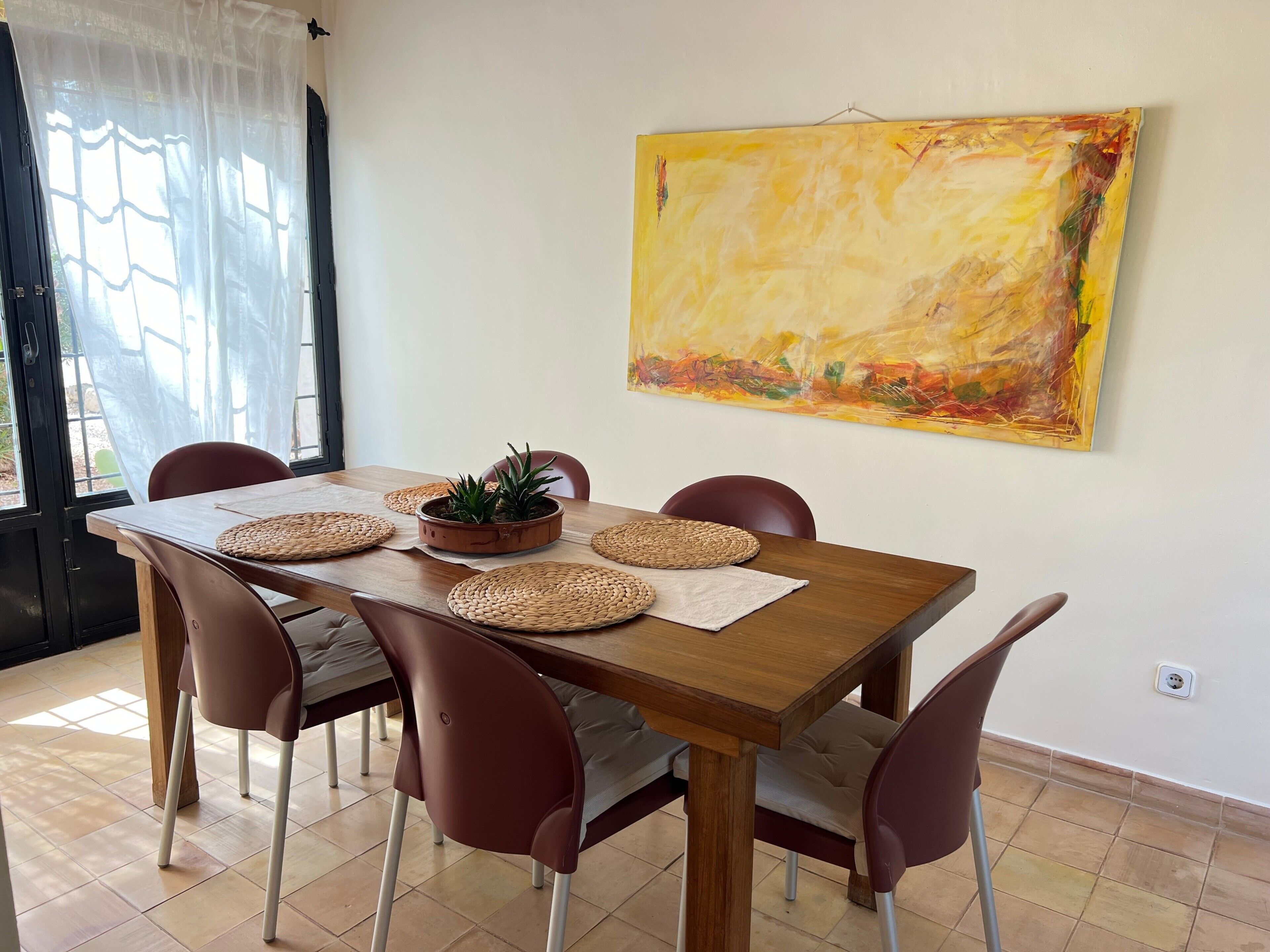 Ein Esstisch mit gewebten Platzsets, einer Topfpflanze und einem modernen Gemälde an der Wand.