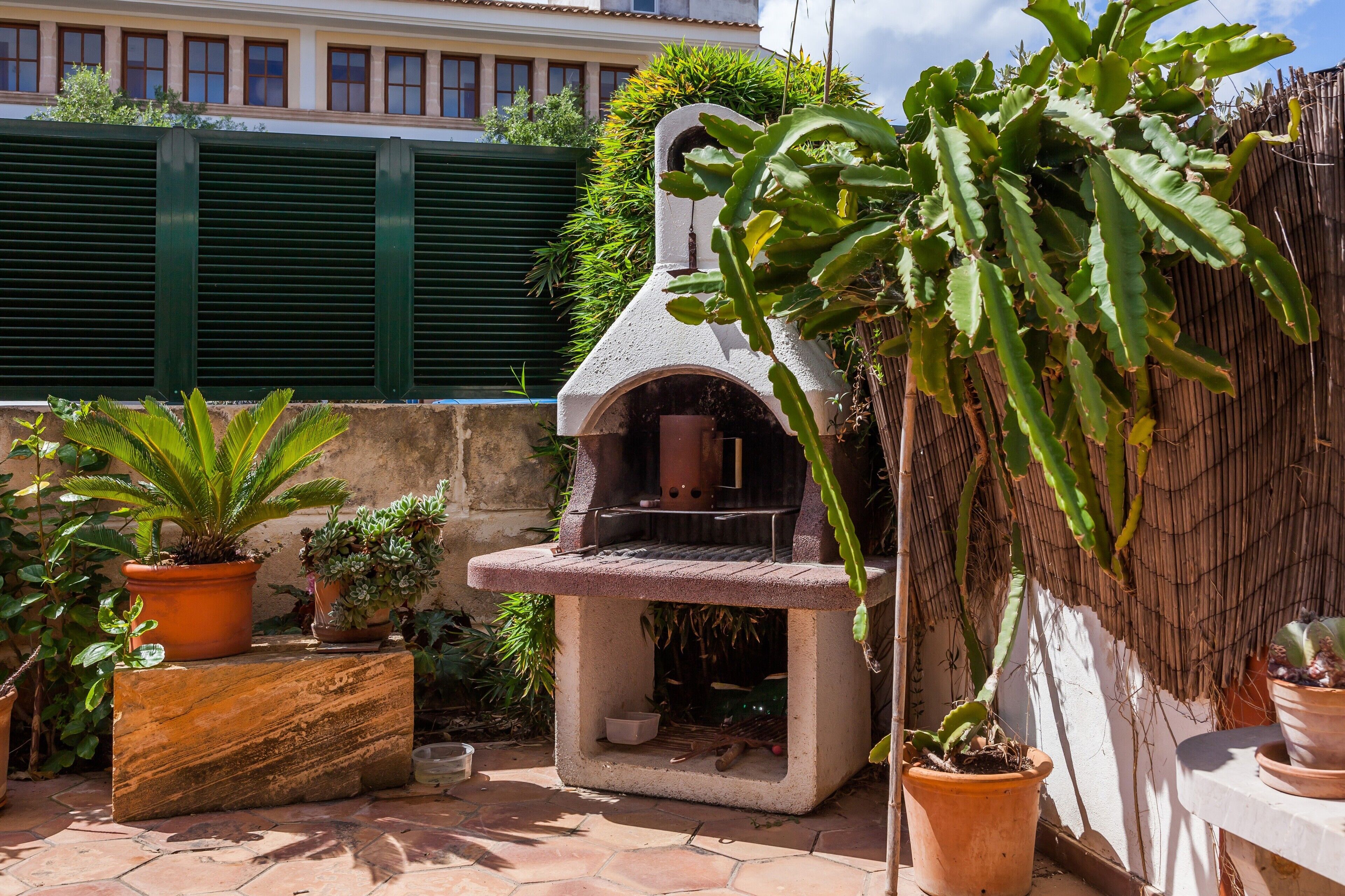 Een terras buiten met een barbecue van steen, planten in potten en een stenen bank.