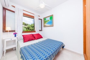 A bedroom with a bed, a nightstand, a lamp, a window with curtains, a painting, and a ceiling fan.