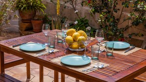 Mesa al aire libre dispuesta con platos, cubiertos y vasos, rodeada de macetas con plantas y un muro de piedra.