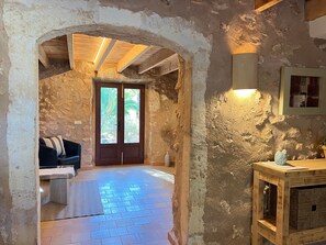 Un intérieur rustique en pierre, avec une porte en bois, un banc rembourré et une petite table agrémentée d’un objet décoratif.