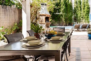 An outdoor dining area with a table set for four, surrounded by chairs, a barbecue grill, and a pool in the background. - Vacation Home "Molino Son Moll" with Pool, Wi-Fi, Balcony, Garden & Terrace (Cala Ratjada)