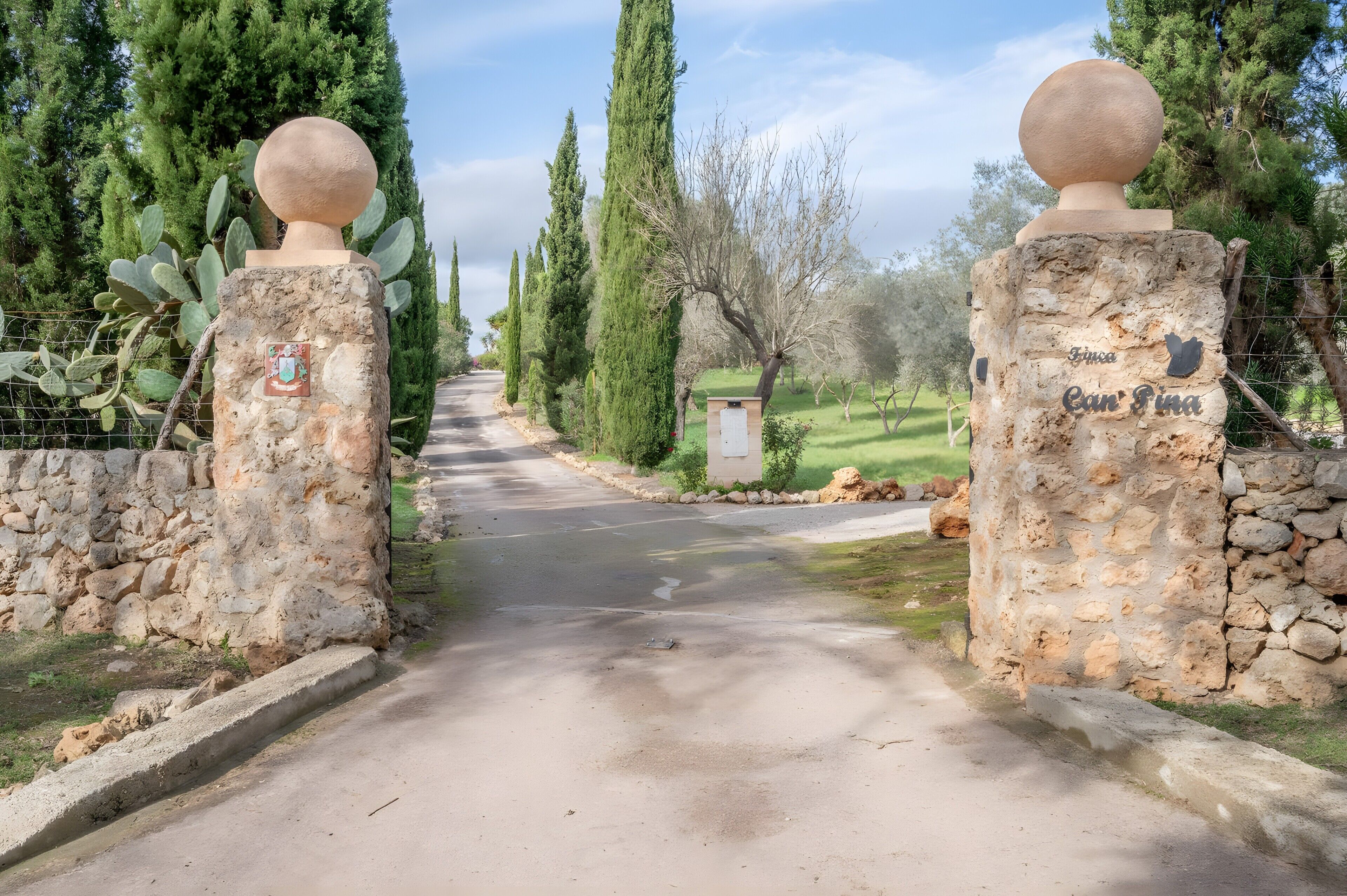A stone pathway leading to a rustic gate with a spherical finial, surrounded by cypress trees and a small building labeled 'Finca Can Fina.'