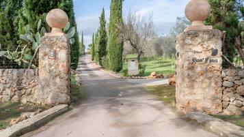 A stone pathway leading to a rustic gate with a spherical finial, surrounded by cypress trees and a small building labeled 'Finca Can Fina.'
