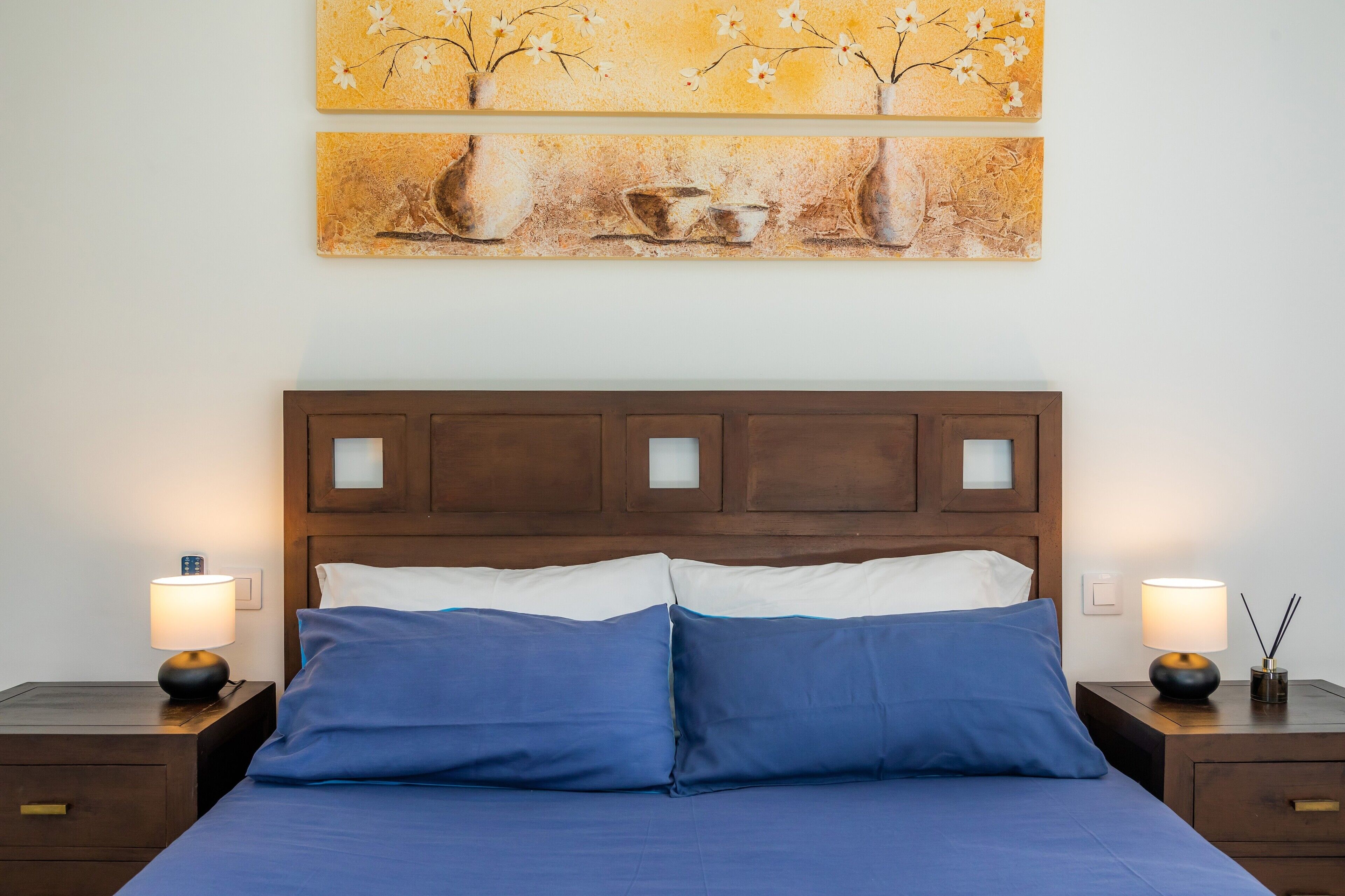 A neatly made bed with blue bedding, two bedside lamps, and two wall-mounted paintings.