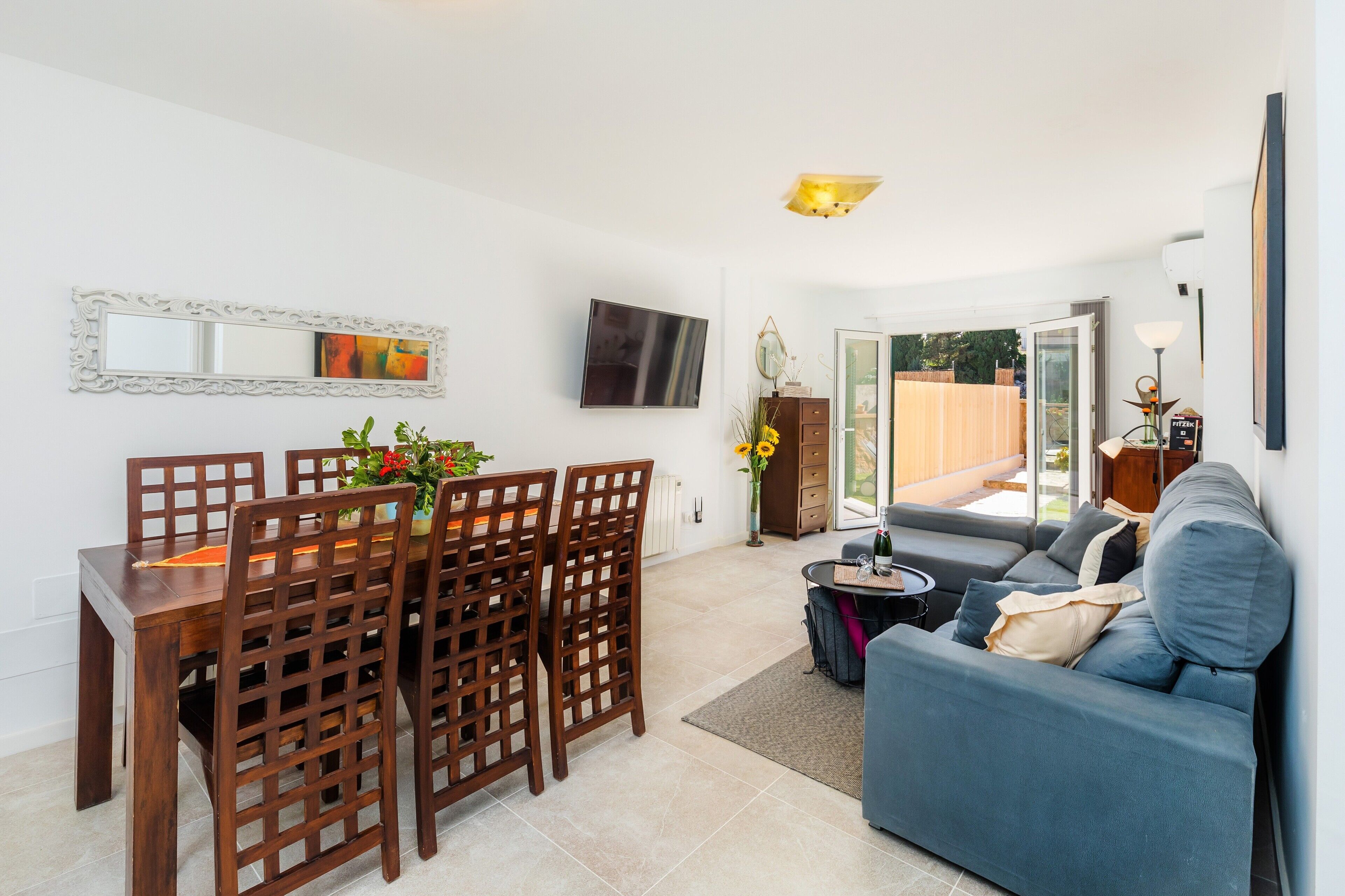 A living room with a sofa, a dining table with chairs, a TV mounted on the wall, and a sliding glass door leading to an outdoor area.
