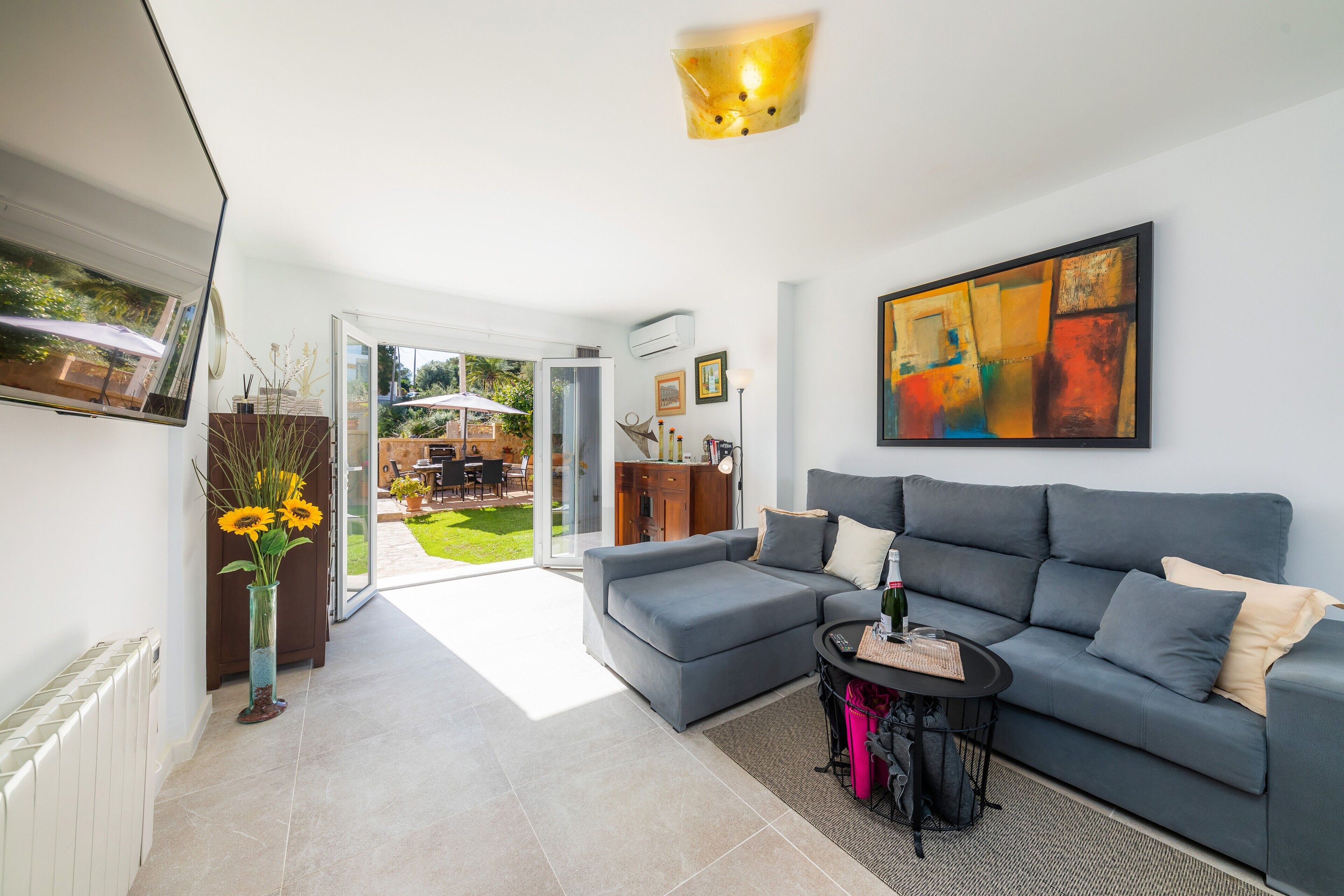 Un salon moderne avec un canapé gris, une table basse et un vase rempli de fleurs. Un tableau orne le mur et une porte donne accès à un espace extérieur.