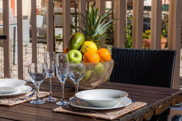 A table with a fruit bowl, wine glasses, and plates, set on an outdoor patio with wicker furniture.