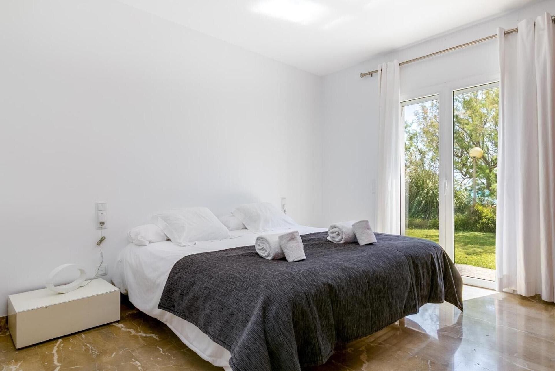 Une chambre à coucher avec un lit, une table de chevet, une lampe et une vue sur l’extérieur.