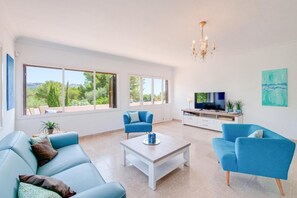 A spacious living room with a large window, a chandelier, a TV, and blue furniture. - Villa Albatros near the Beach, with Sea View, Garden, Wi-Fi and A/C (Canyamel)