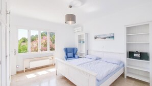 A bedroom with a bed, a chair, a bench, a bookshelf, and a window with a view of greenery. - Villa Albatros near the Beach, with Sea View, Garden, Wi-Fi and A/C (Canyamel)