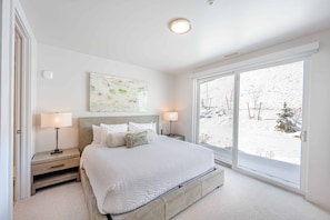 Ein Schlafzimmer mit einem großen Bett, zwei Nachttischen, einer Stehlampe, einer Deckenleuchte, einem Fenster mit Blick auf eine verschneite Landschaft und einem abstrakten Kunstwerk an der Wand.