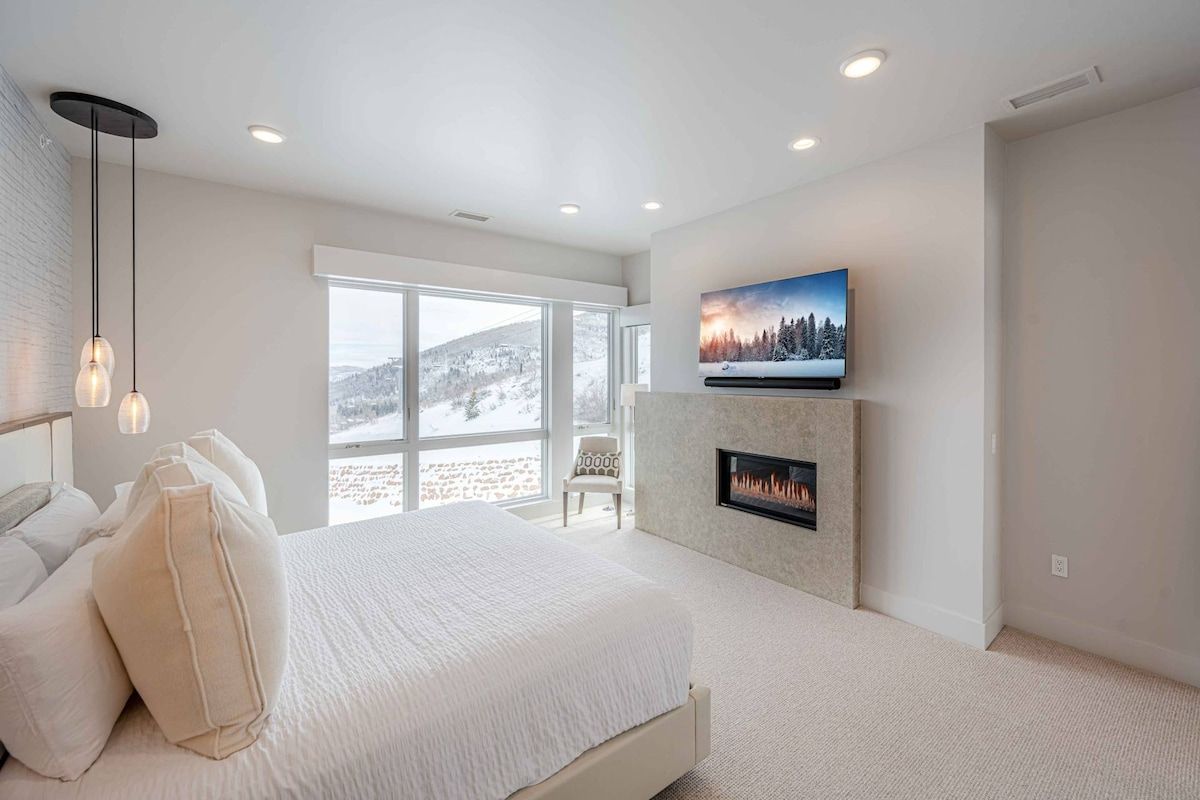 A bedroom with a large bed, a fireplace, a television, and a view of a snowy landscape through large windows.