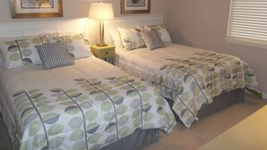Ein Schlafzimmer mit zwei Betten, jeweils mit blattgemusterten Bettbezügen, einer Nachttischlampe und gerahmten Bildern an der Wand.