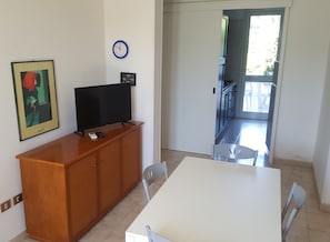 A room with a wooden cabinet, a flat-screen TV, a clock, a framed picture, and a table with chairs.