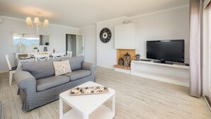Ein modernes Wohnzimmer mit grauem Sofa, weißem Couchtisch, Flachbildfernseher, Kamin und Essbereich mit weißem Tisch und Stühlen.