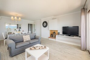 A modern living room with a grey sofa, a white coffee table, a flat-screen TV, a fireplace, and a dining area with a white table and chairs.