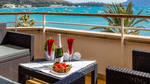 Ein Balkon mit Korbmöbeln, einem Tisch mit Flasche, Gläsern und Erdbeeren, mit Blick auf einen Yachthafen mit Booten und Hügeln im Hintergrund.