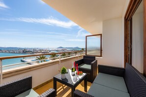 A balcony with wicker furniture, a table with a bottle and glass, and a view of the marina and mountains.
