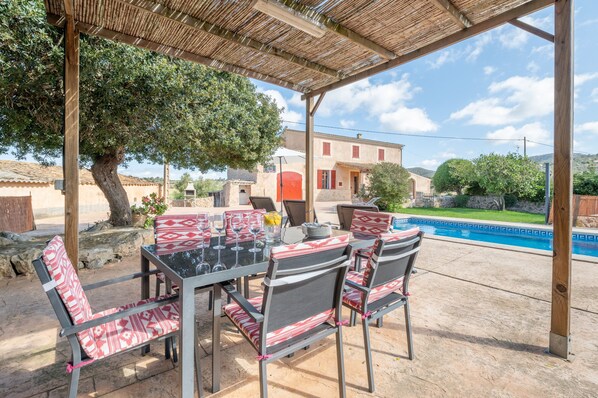 An outdoor patio with a table and chairs, a pool in the background, and a building with a red door.