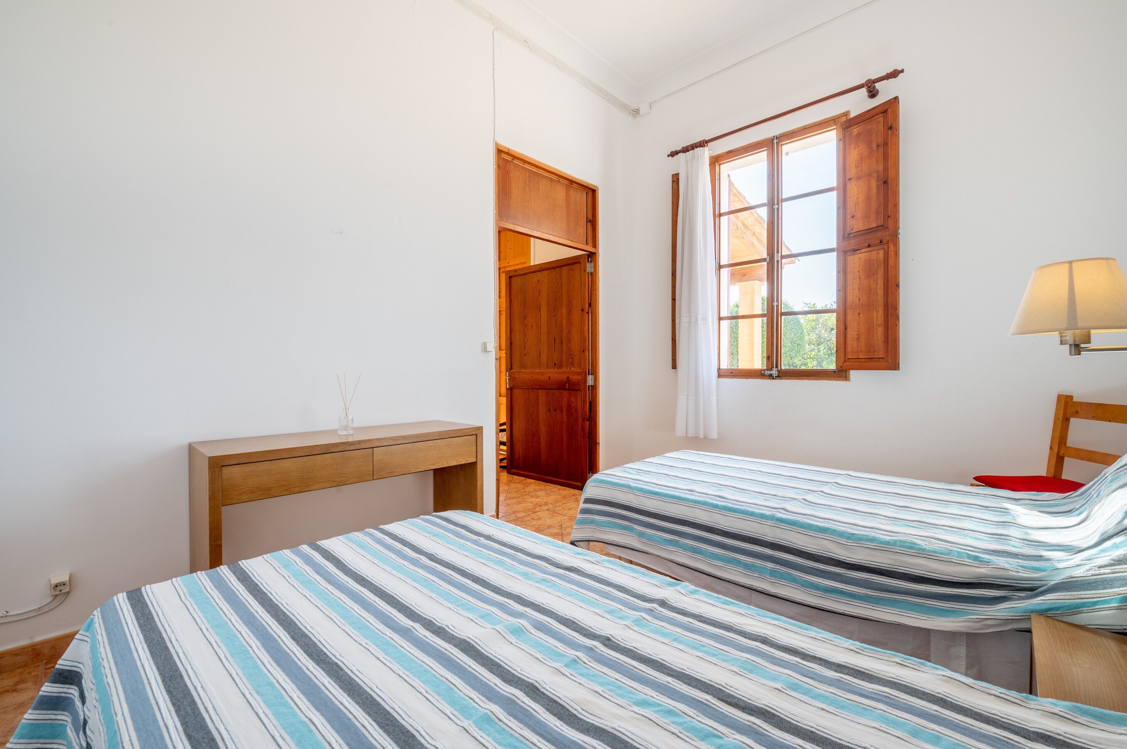A bedroom with two beds, a wooden desk, a window with curtains, and a door leading to another room.
