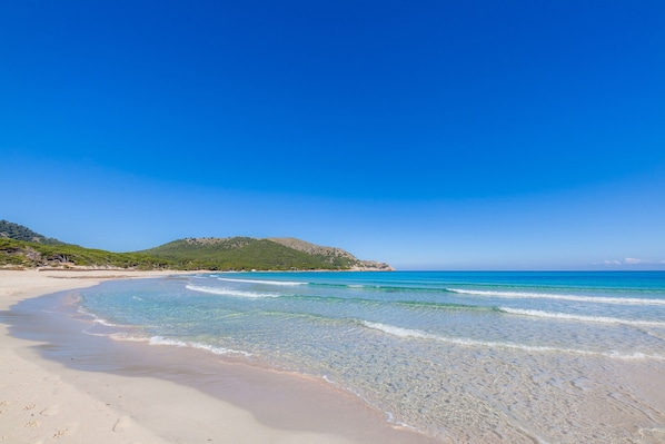 A beach with clear water, a sandy shore, and a green hill in the background.