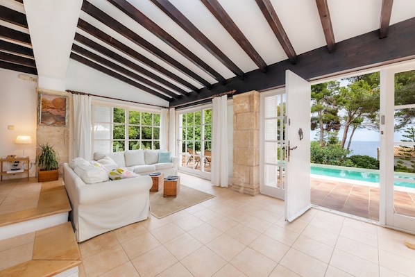 A spacious living room with a white sofa, a coffee table, and a view of a pool and greenery.