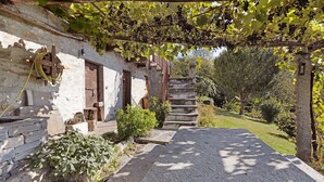 Un bâtiment rustique en pierre, doté d’un escalier en bois menant à une porte, est entouré de verdure et d’une pergola couverte de vignes.